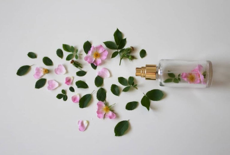 A glass perfume bottle with a gold sprayer lies on its side, surrounded by scattered pink flower petals and green leaves on a white background.