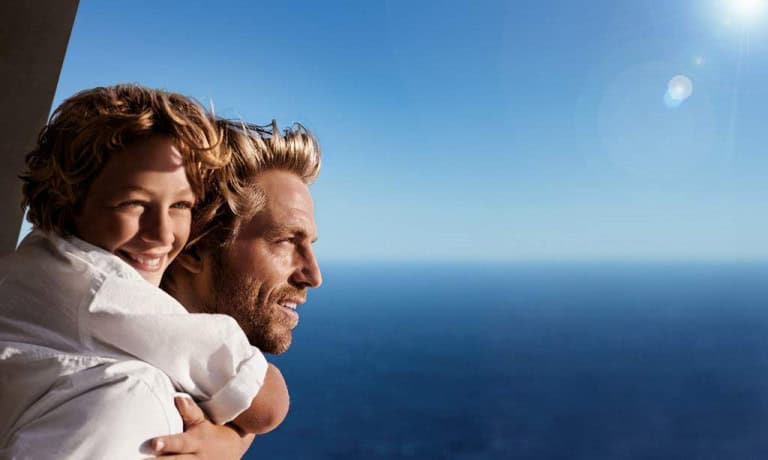 A smiling child hugs a man from behind as they look out over a vast, blue ocean under a clear sky with bright sunlight. Both appear happy and relaxed.