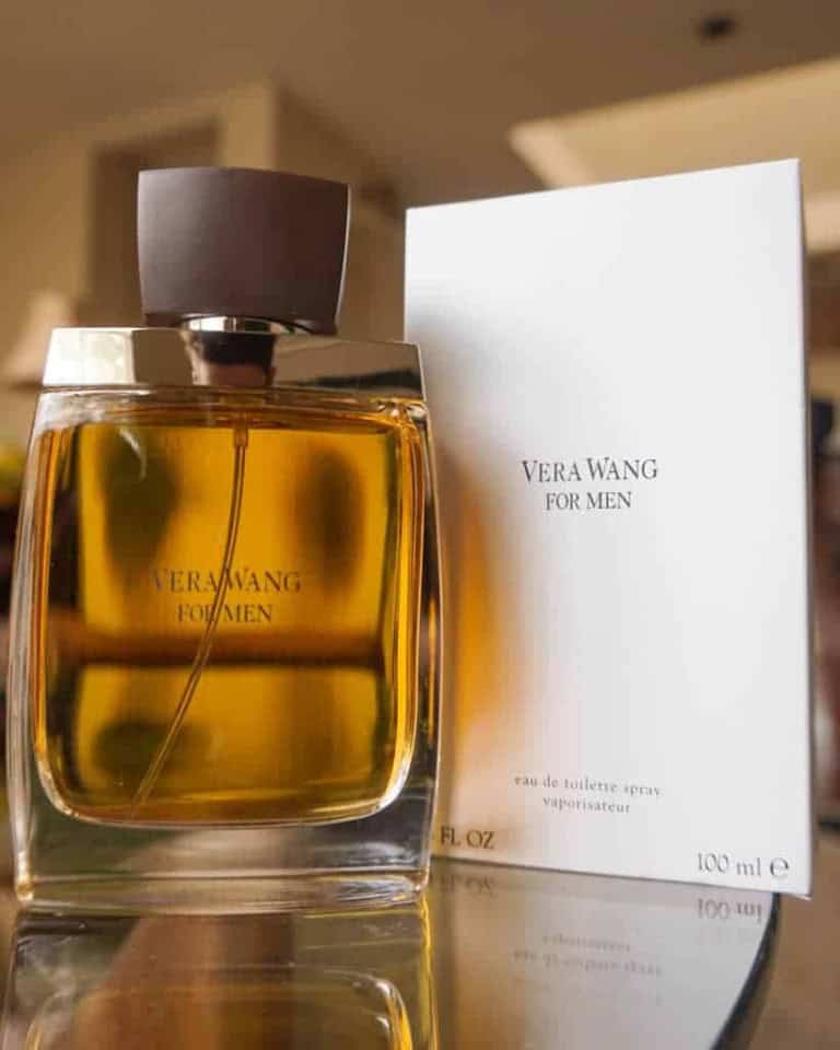 A bottle of Vera Wang For Men eau de toilette sits next to its white packaging box on a reflective surface, with a blurred indoor background. The bottle contains amber-colored liquid.