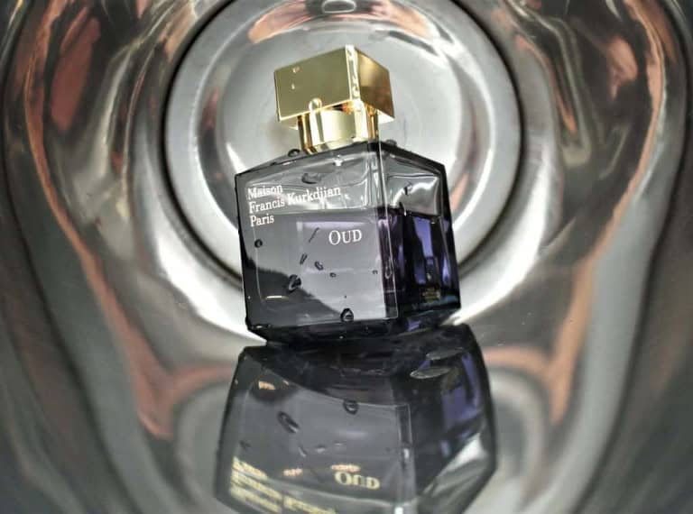 A bottle of Maison Francis Kurkdjian Paris Oud perfume with a gold cap is positioned upright, reflected against a shiny metallic or mirrored surface.