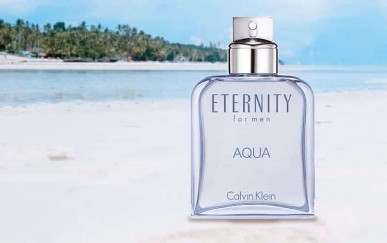 A bottle of Calvin Klein Eternity for Men Aqua cologne is positioned on a sandy beach with the sea and palm trees in the background under a clear blue sky.