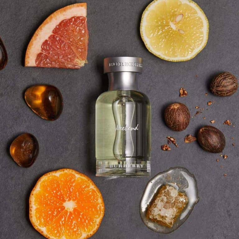 A bottle of Burberry Weekend perfume is surrounded by slices of grapefruit, lemon, and orange, whole nutmegs, amber stones, and a piece of honeycomb on a dark surface.