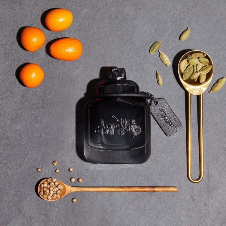 A black perfume bottle with a Coach tag is placed on a gray surface, surrounded by whole kumquats, a golden measuring spoon with cardamom pods, a wooden spoon with coriander seeds, and loose cardamom pods.