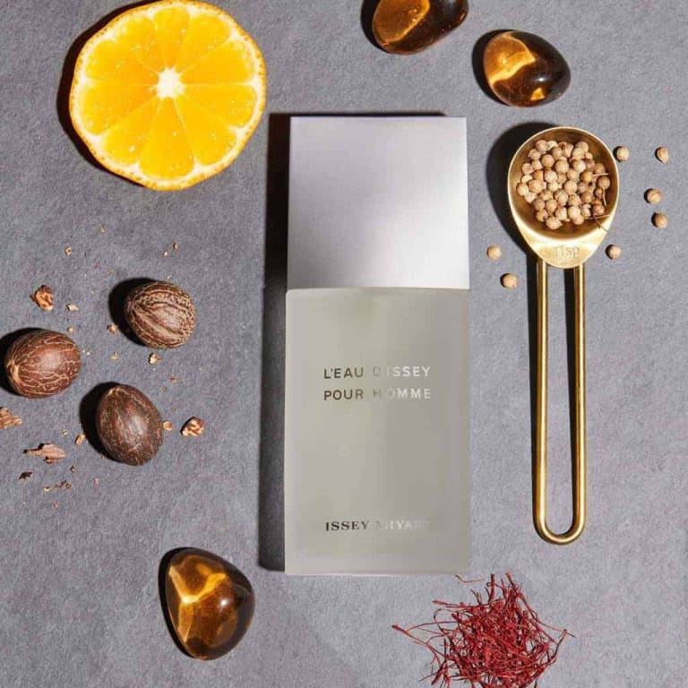 A bottle of LEau dIssey Pour Homme perfume is placed on a dark surface, surrounded by half an orange, nutmeg, saffron threads, a gold spoon with peppercorns, and amber stones.