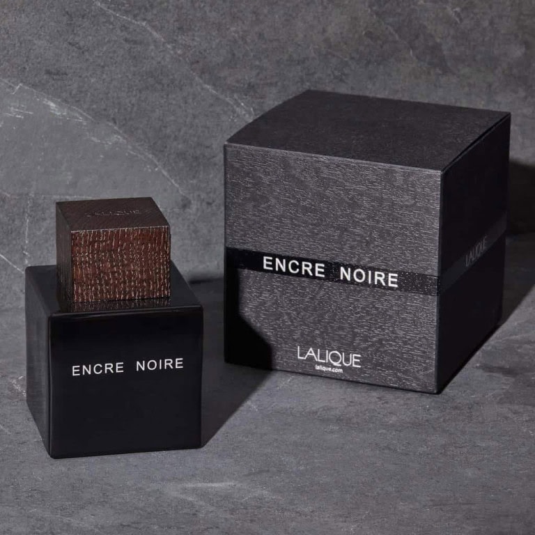 A black bottle of Lalique Encre Noire perfume with a textured brown cap is placed next to its matching black box, both sitting on a dark grey stone surface.