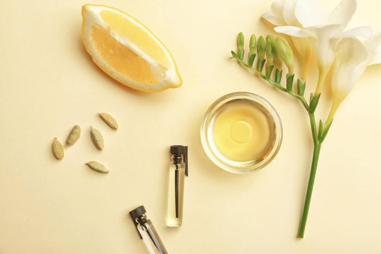 A lemon wedge, a white flower, green buds, cardamom pods, a small bowl of oil, and two small glass vials with black caps are arranged on a pale yellow background.
