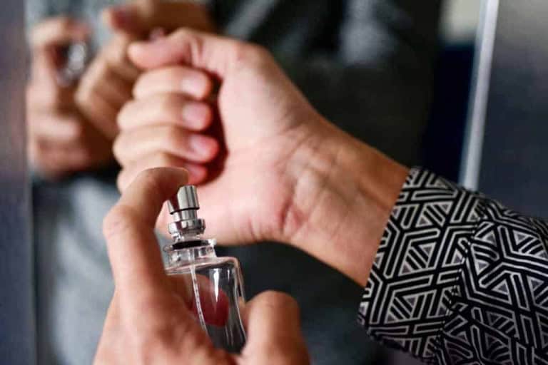 A person sprays perfume from a clear bottle onto their wrist, wearing a shirt with a black and white geometric pattern sleeve, with their reflection visible in a mirror.
