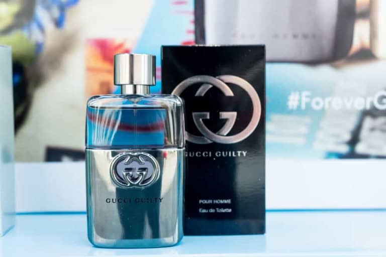 A bottle of Gucci Guilty Pour Homme eau de toilette is displayed next to its black packaging box, both featuring the Gucci logo, against a blurred background with blue and white colors.
