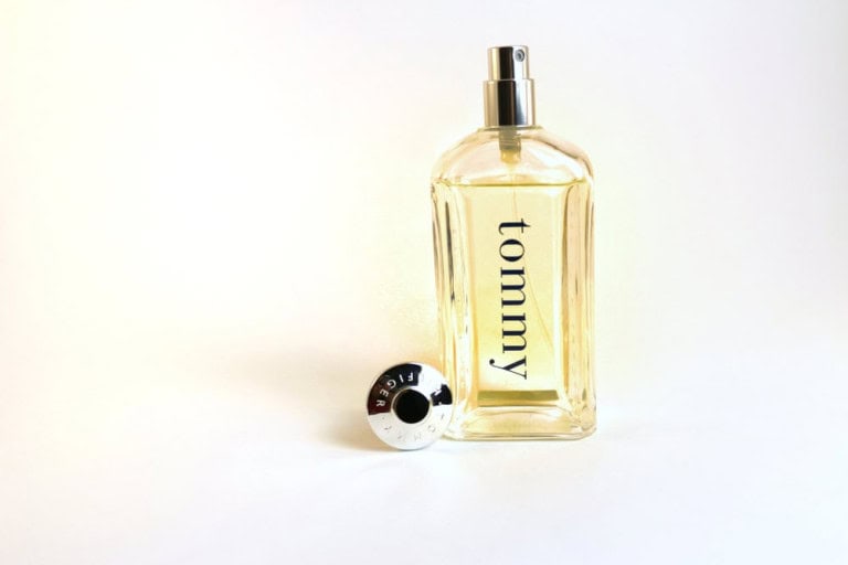 A clear glass bottle of Tommy Hilfiger perfume with a silver cap placed beside it on a white background. The bottle contains a yellowish liquid and the word tommy is printed vertically on the front.