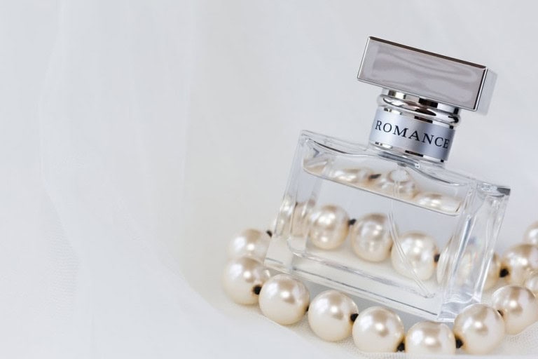 A glass perfume bottle labeled Romance is surrounded by a string of white pearls, resting on a soft white fabric background.