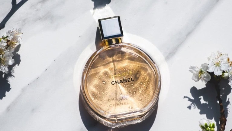 A round bottle of Chanel Chance perfume sits on a white marble surface, with light shining through the bottle and casting a shadow. White flowers surround the perfume, creating a fresh, elegant scene.