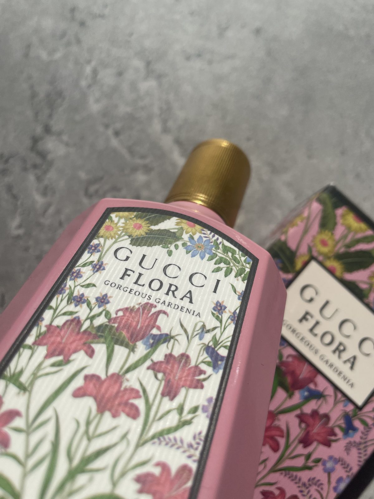 Gucci Flora Gorgeous Gardenia Review: The Flower That Isn't There Gucci Flora Gorgeous Gardenia bottle in focus with its pink floral box behind on concrete