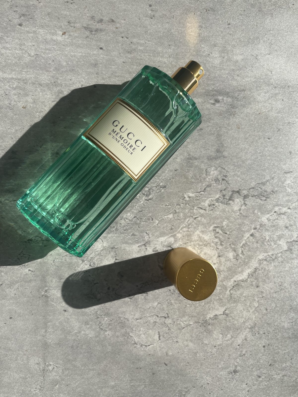 Gucci Memoire d’une Odeur EDP Review: Soothing & Soft Gucci Memoire d’une Odeur EDP bottle lying on concrete in direct sunlight with the gold cap beside it