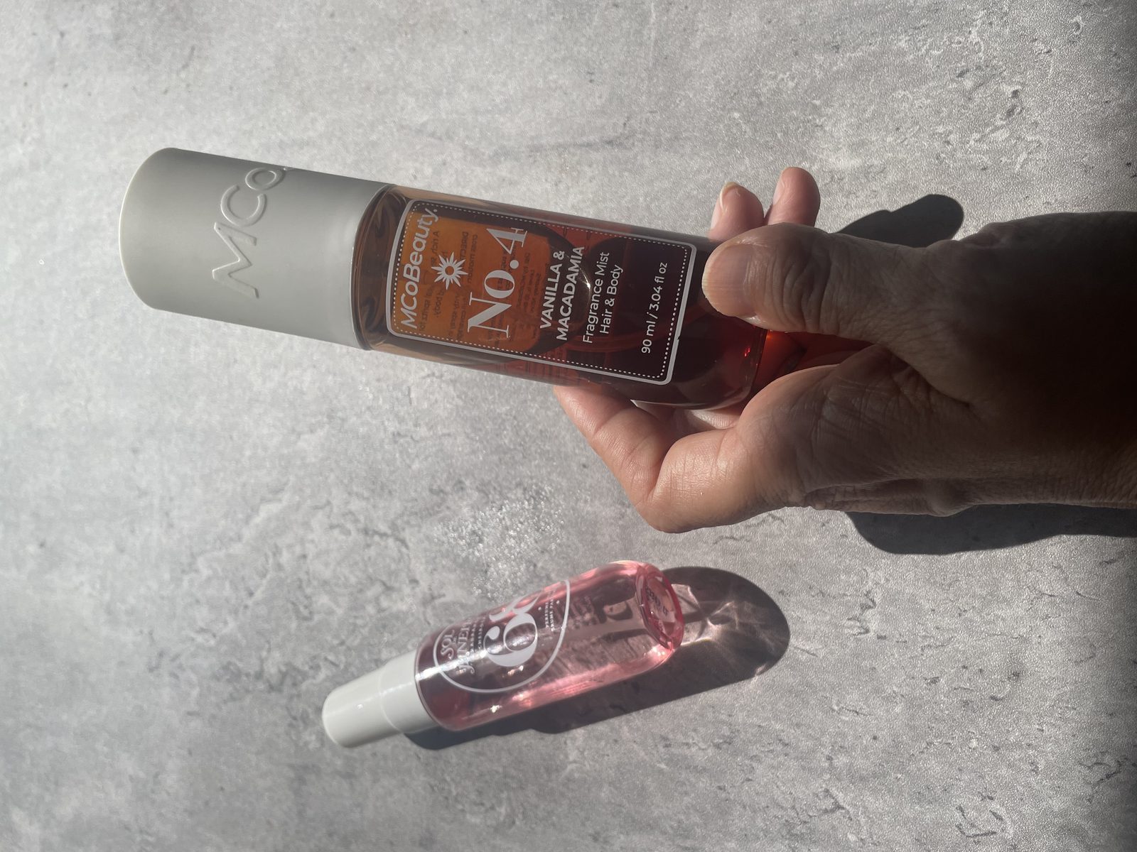 MCoBeauty vs Sol de Janeiro: I Own Both, and They Don't Smell Alike Holding MCoBeauty No.4 Vanilla & Macadamia with Sol de Janeiro Cheirosa 68 on the concrete below