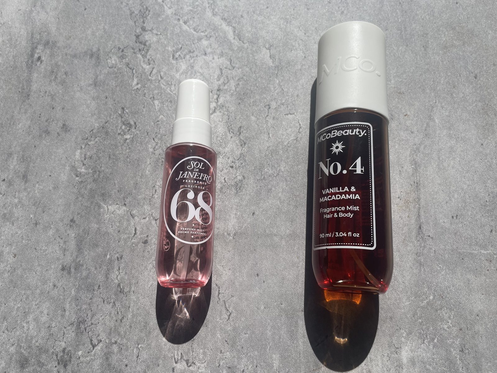 MCoBeauty vs Sol de Janeiro: I Own Both, and They Don't Smell Alike MCoBeauty No.4 Vanilla & Macadamia and Sol de Janeiro Cheirosa 68 standing side by side on sunlit concrete, both labels fully readable