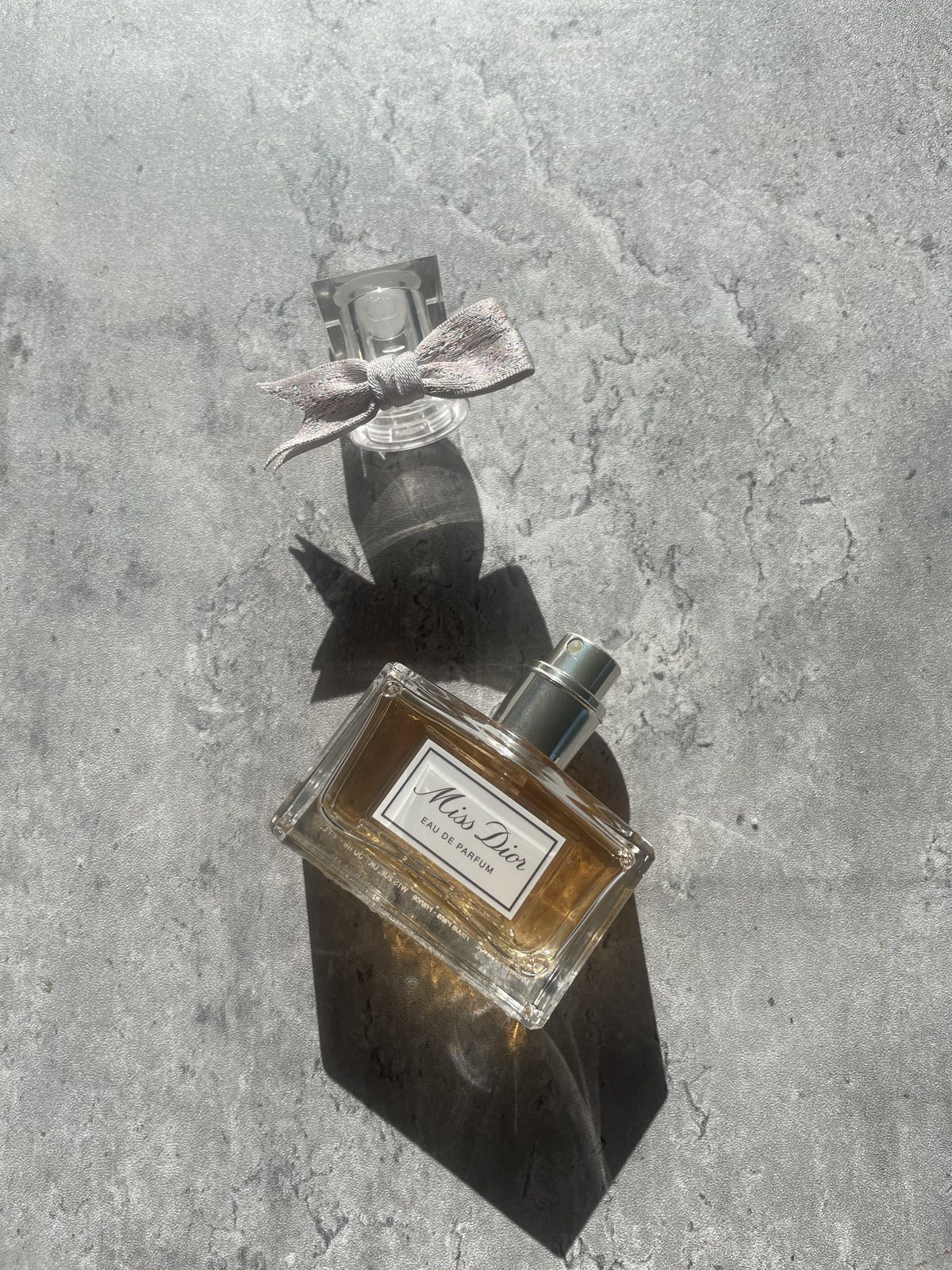 Miss Dior Perfume: The Modern and Airy Fragrance Reviewed Miss Dior Eau de Parfum 30ml bottle on sunlit concrete with the bow-tied cap beside it