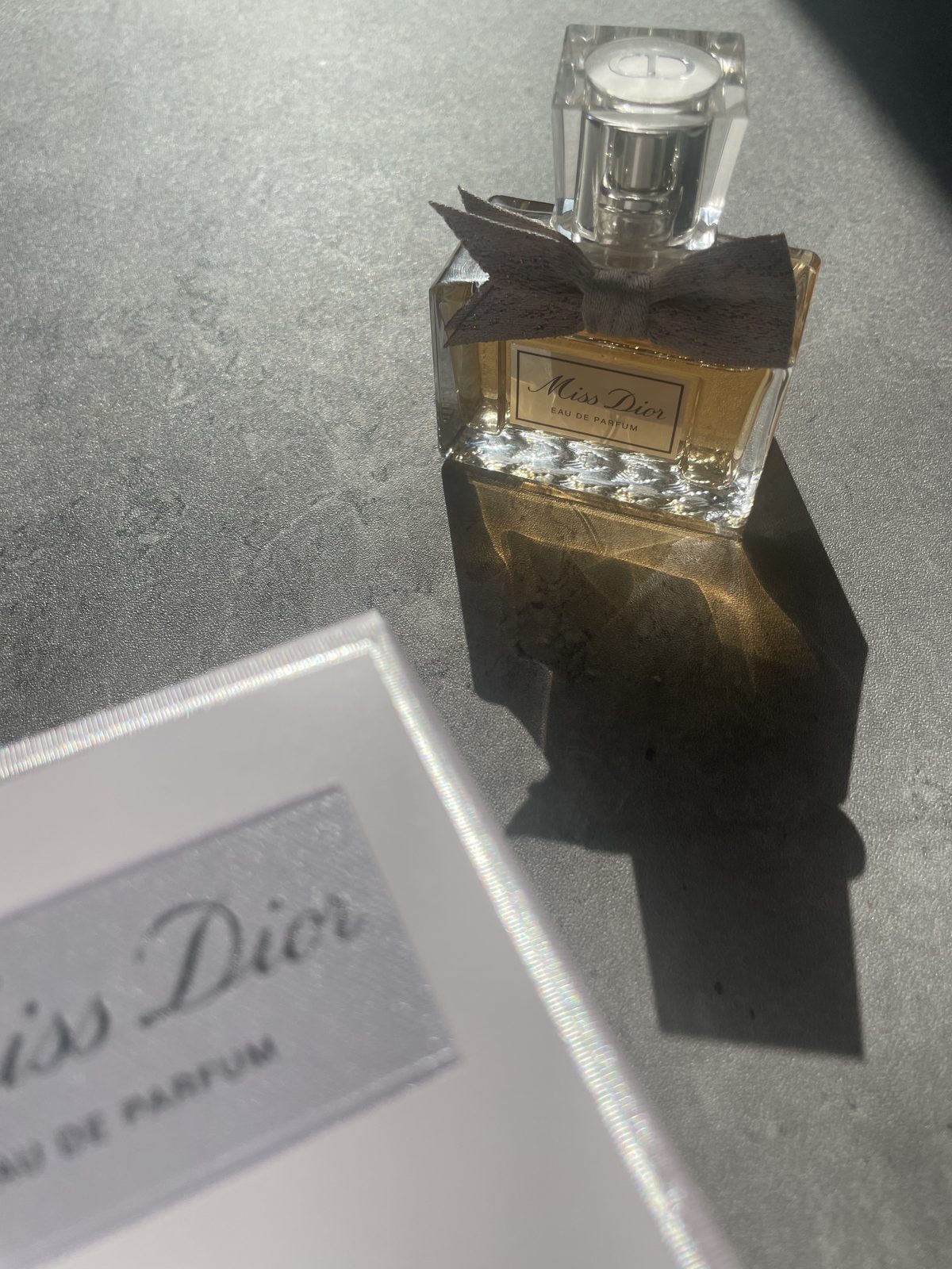 Miss Dior Perfume: The Modern and Airy Fragrance Reviewed Miss Dior Eau de Parfum bottle standing upright with the bow and Dior medallion cap, box beside it on concrete