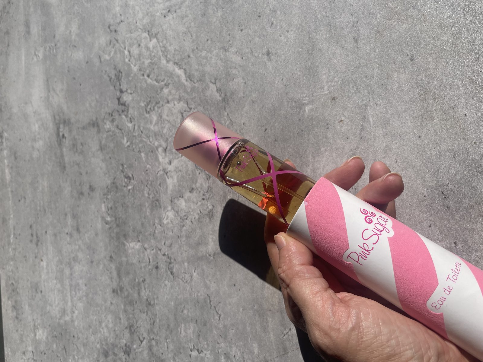 Pink Sugar Perfume Reviewed: Incredibly Sweet Holding the Aquolina Pink Sugar Eau de Toilette bottle sliding out of its striped tube, on sunlit concrete